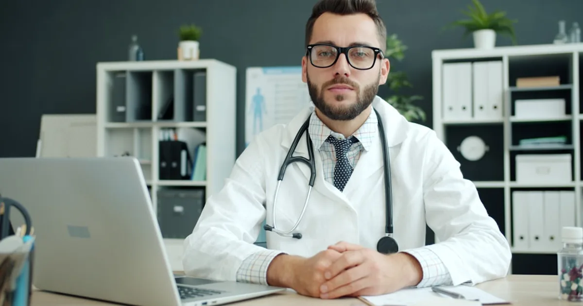 Ärztin in weißem Kittel sitzt an einem Schreibtisch mit Monitor in einer modernen Arztpraxis.